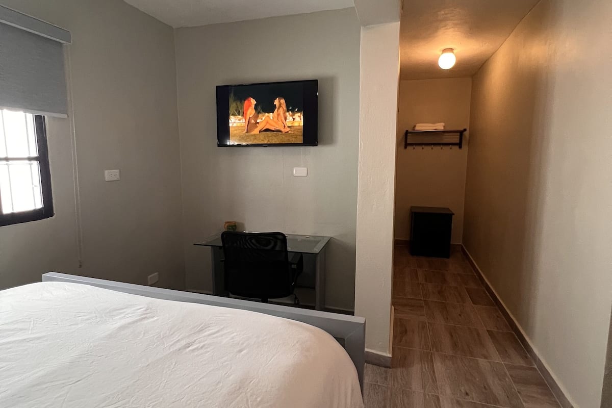 Een slaapkamer met een bed, een bureau, een stoel, een aan de muur bevestigde televisie en een hal met een lamp.