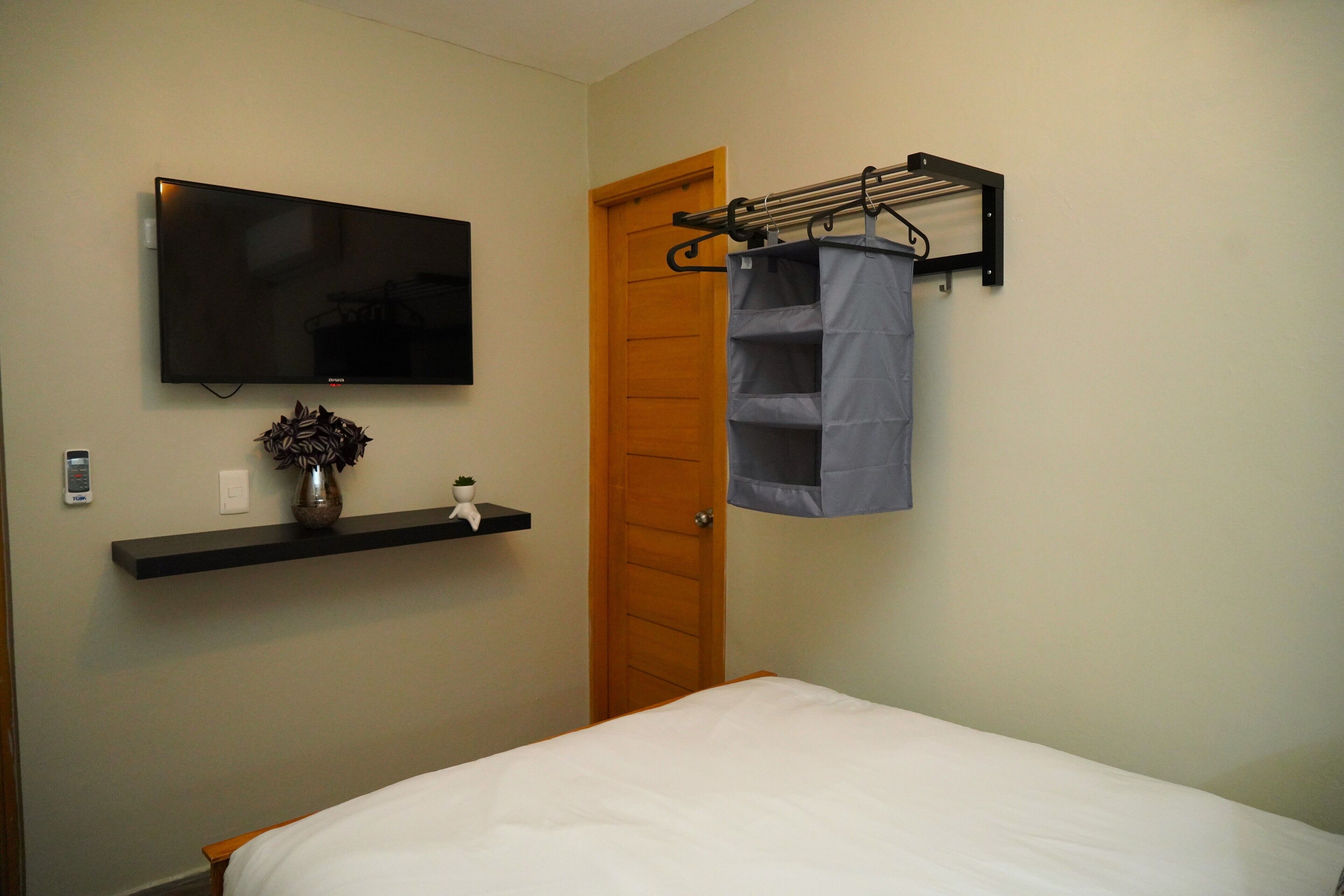 Un dormitorio con cama, televisor montado, estante con un jarrón y una puerta.