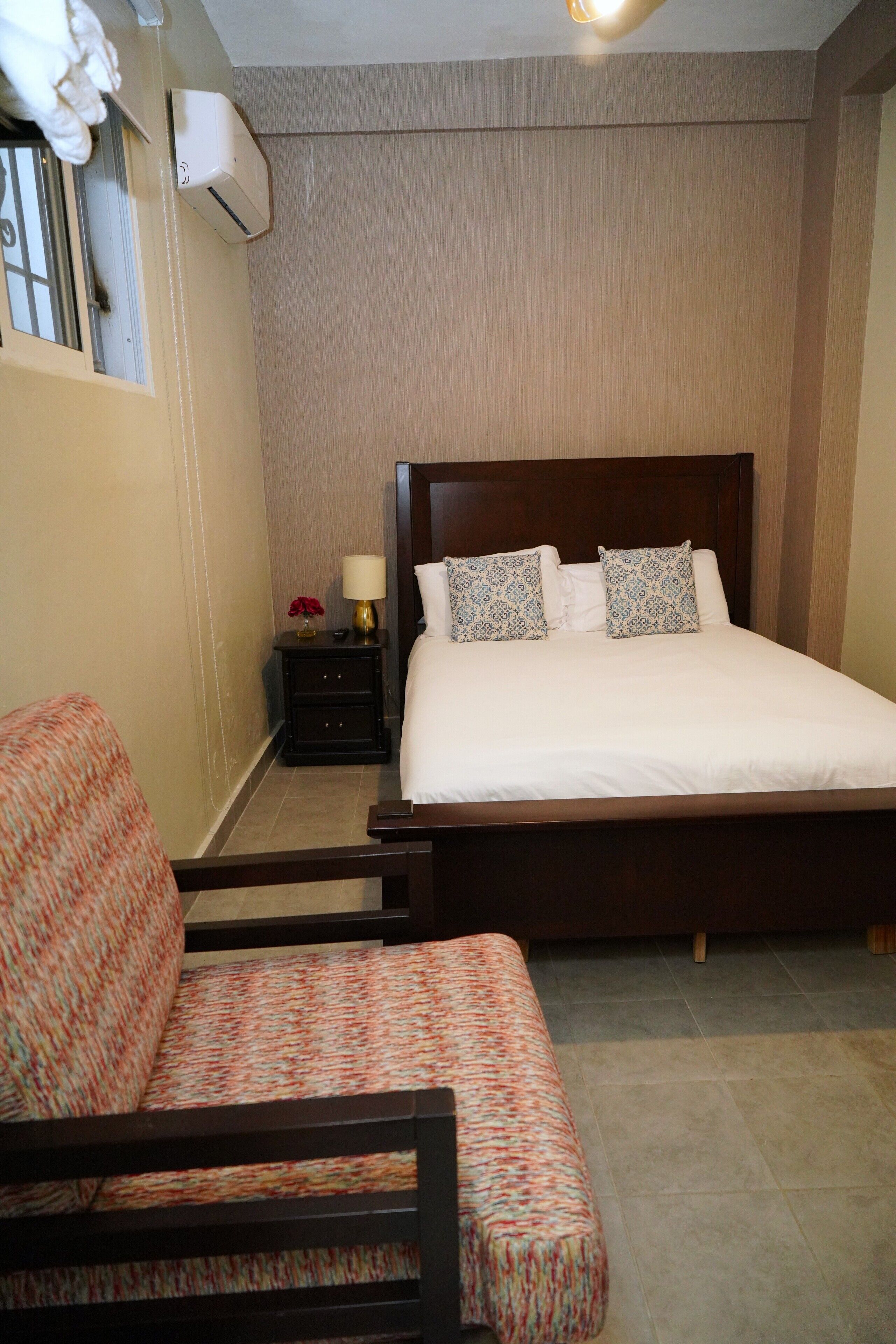 A hotel room with a large bed, a bedside table with a lamp, a chair, and an air conditioning unit on the wall.
