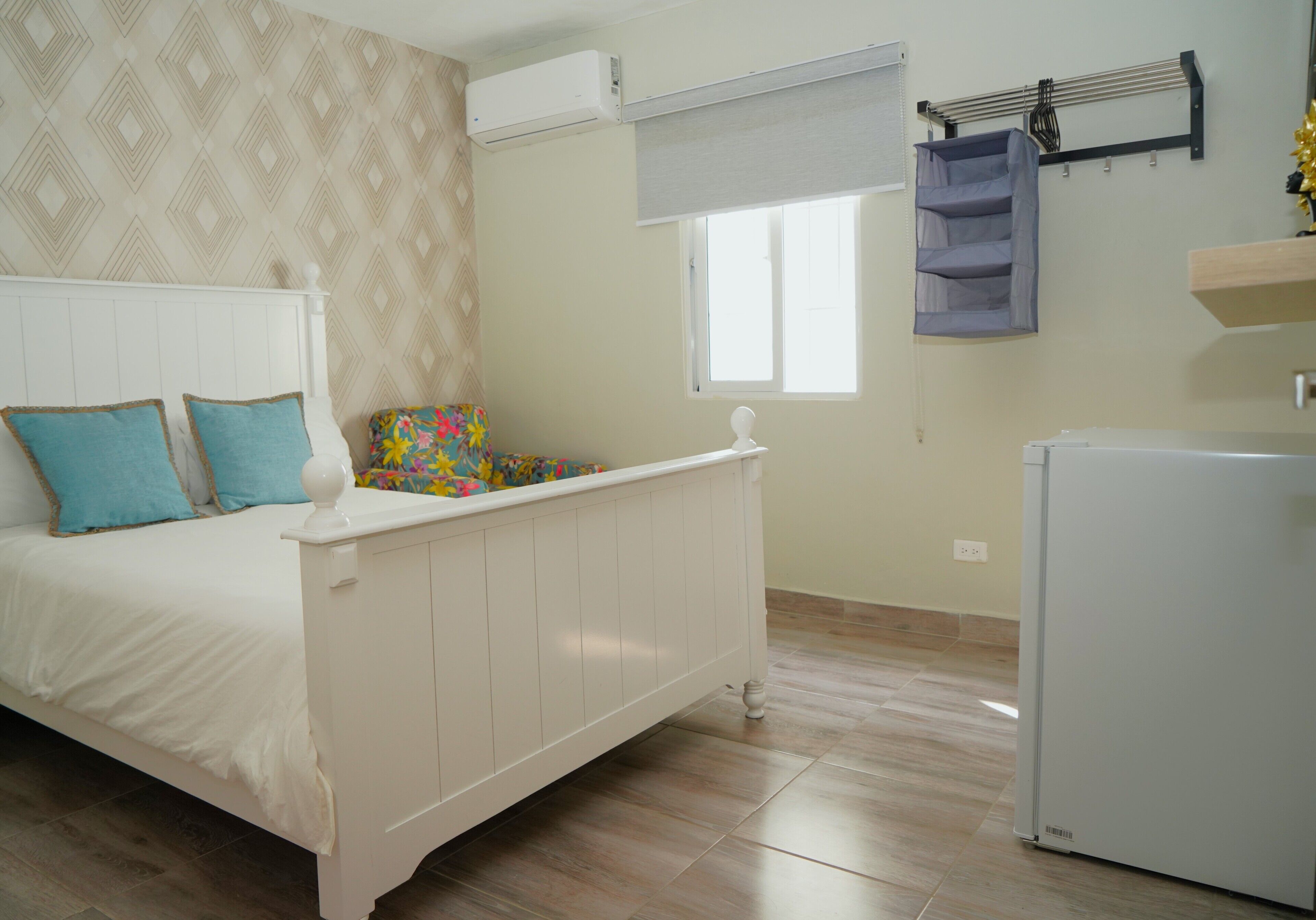 Quarto com cama branca, radiador, janela e frigorífico branco.