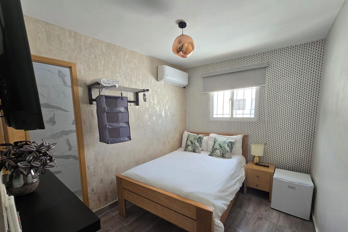 Een slaapkamer met een houten bed, een klein nachtkastje, een wandairconditioner, een raam met versierde gordijnen en een bloempot.