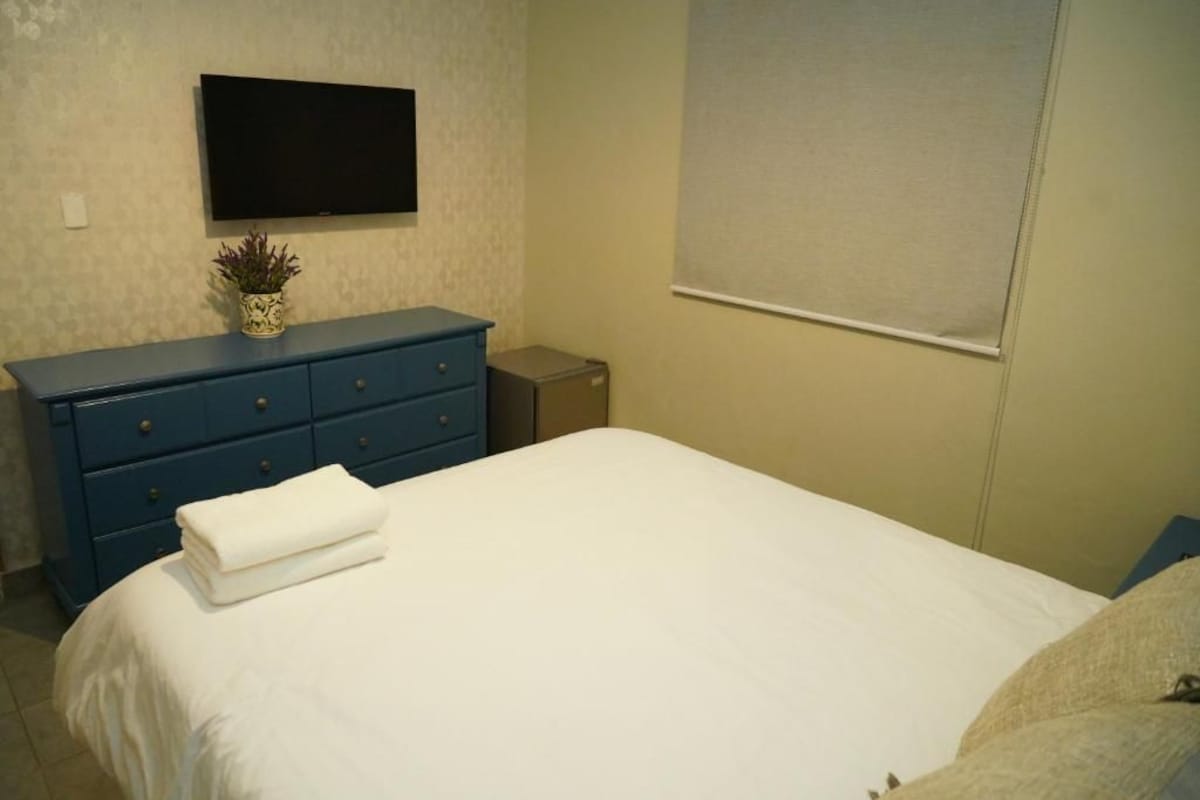 A bedroom with a bed, a dresser, a television mounted on the wall, and a window with blinds.