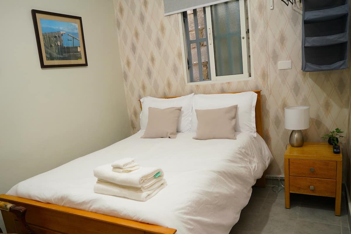 Een netjes opgemaakt bed met witte lakens en een gevouwen handdoek, een nachtkastje met een lamp en een afstandsbediening, en een stoel met een bloemenkussen.