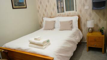 A neatly made bed with white linens and a folded towel, a bedside table with a lamp and a remote control, and a chair with a floral cushion.