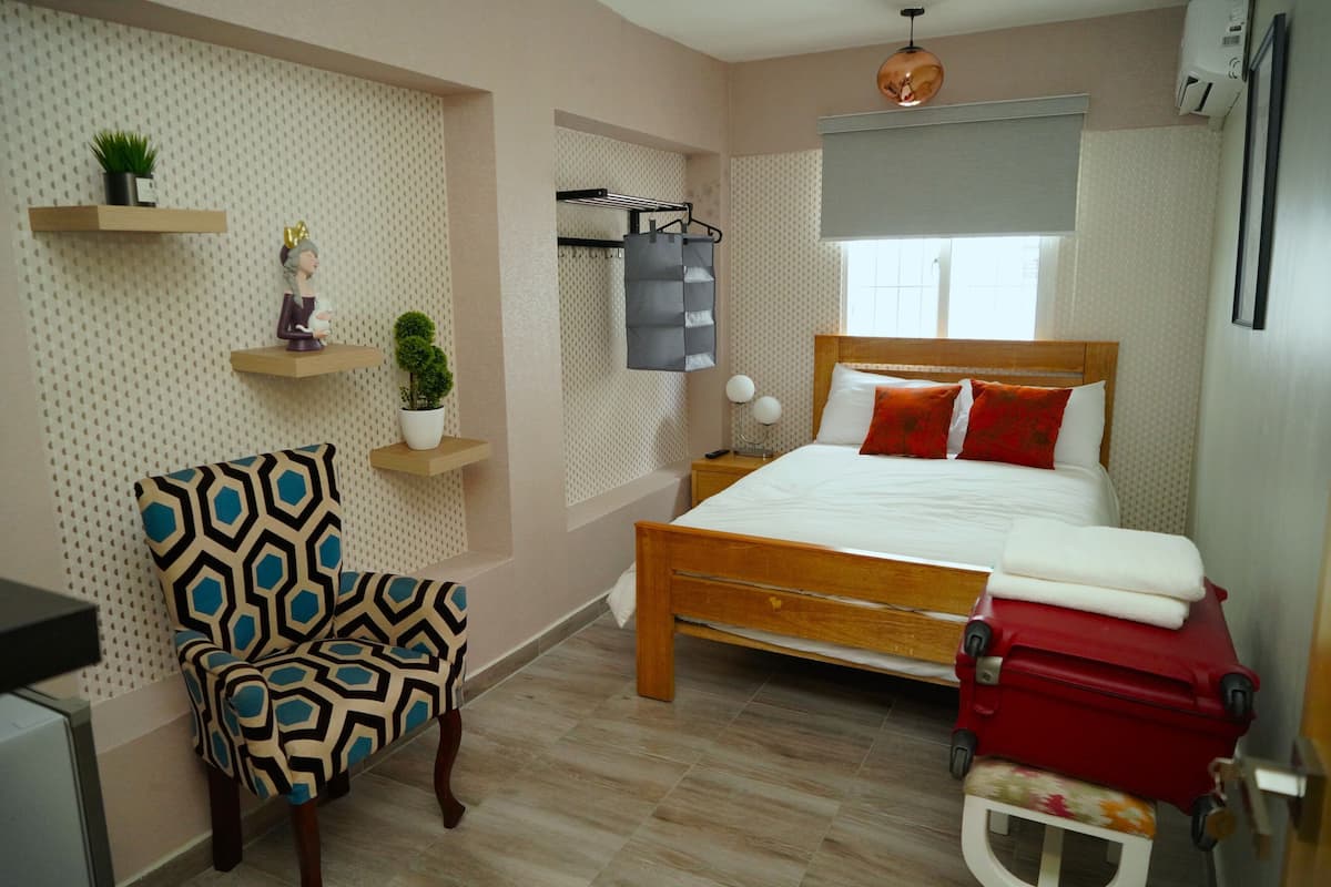 Een slaapkamer met een bed, een stoel, een klein tafeltje, een plank met planten en een koffer.