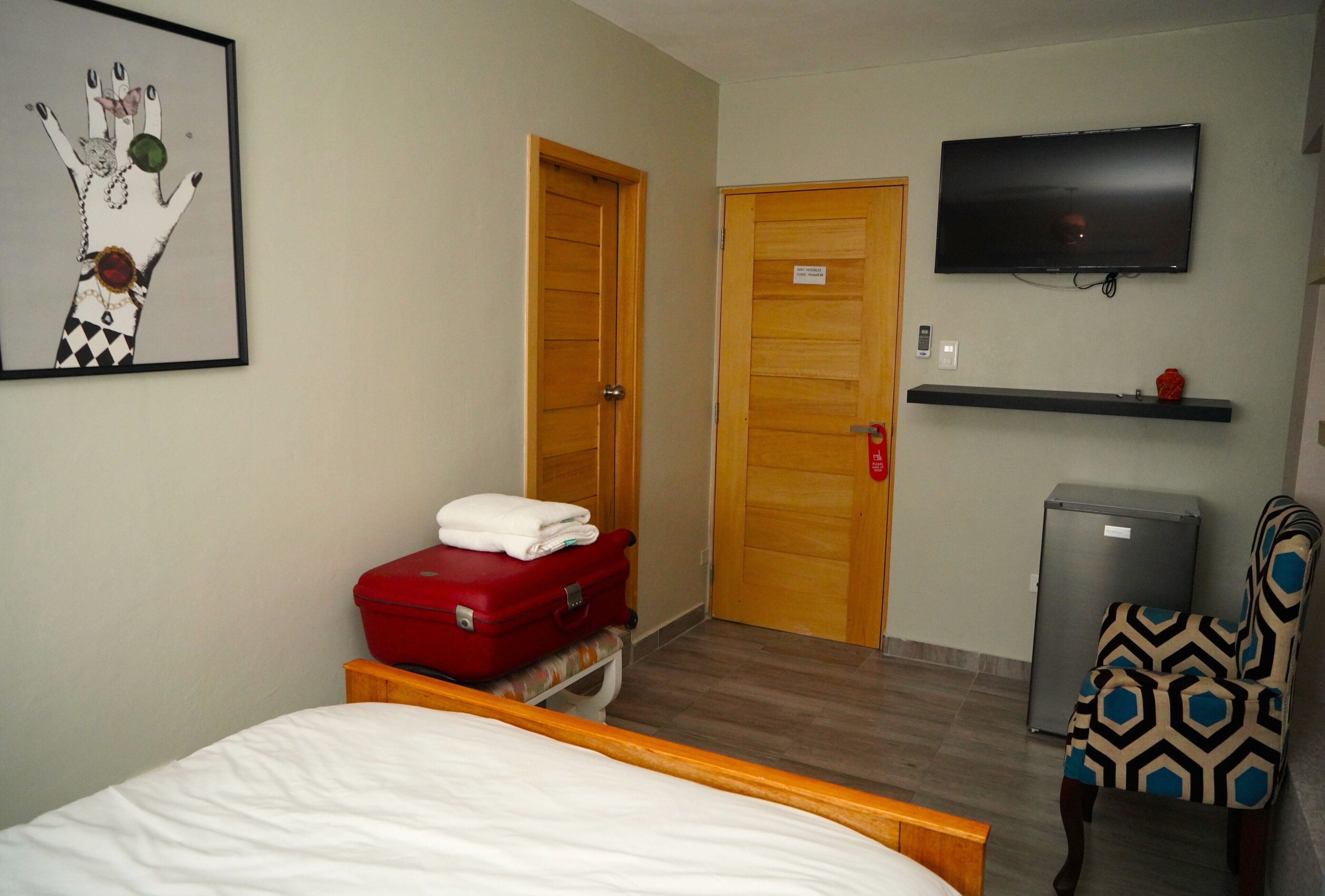 Une chambre d’hôtel avec un lit, une valise rouge, un téléviseur, un petit réfrigérateur et une chaise à motifs.