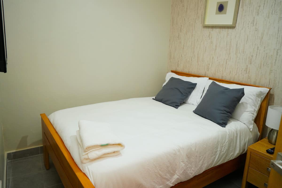 Een netjes opgemaakt bed met witte en grijze linnens, een houten hoofdbord en een klein nachtkastje met een lamp.