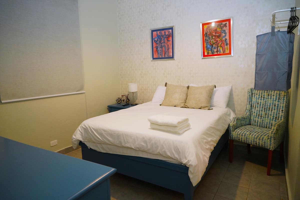 Een slaapkamer met een bed, twee kussens, een stoel, een nachtkastje en twee ingelijste kunstwerken aan de muur.