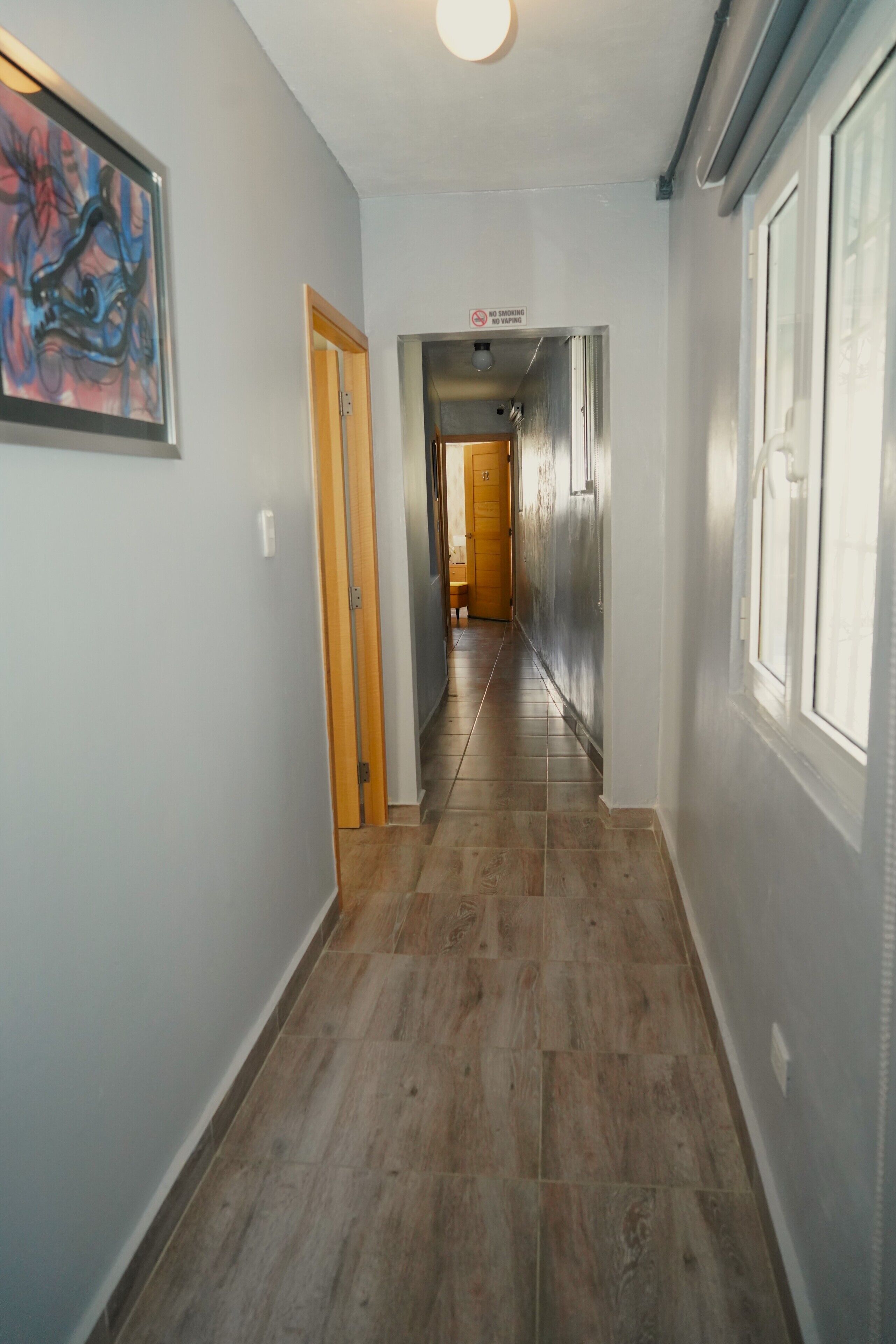 A hallway with a wooden floor, a framed abstract painting on the left, and a door with a 'No Smoking' sign.