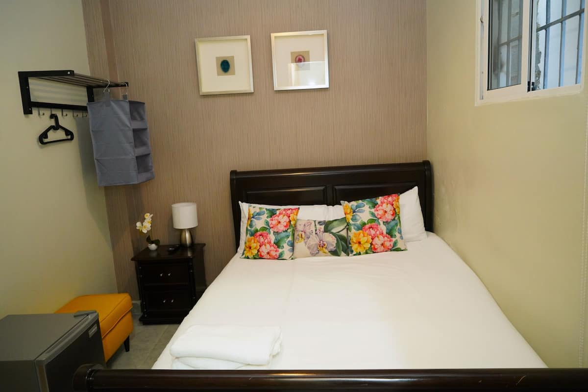 Een net opgemaakt bed met bloemenkussens, een nachtkastje met een lamp en een wandrek voor het drogen van kleding.