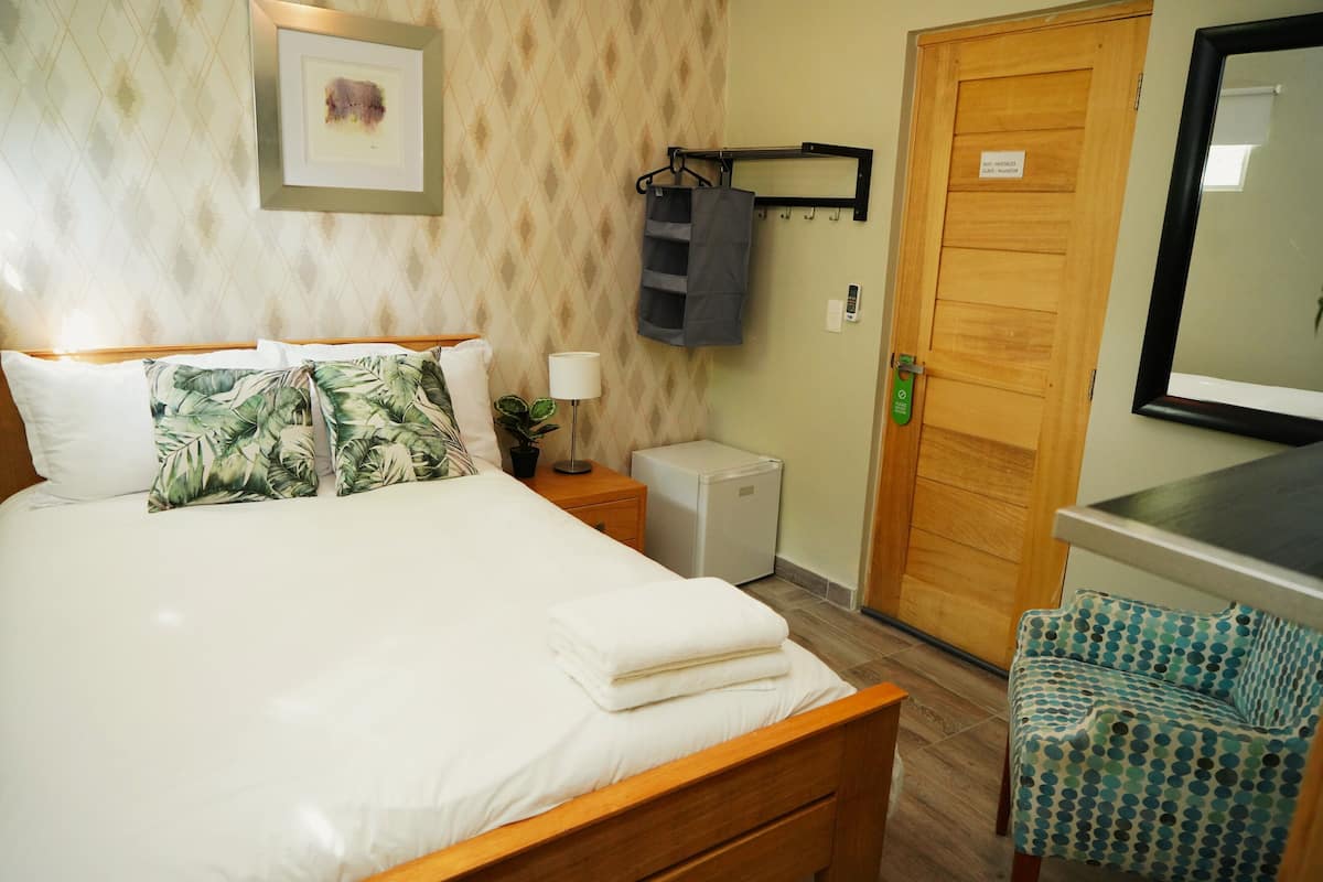 Een net opgemaakt bed met witte lakens en kussens met een tropisch patroon, een nachtkastje met een lamp, een kleine koelkast, een houten deur met een bordje en een spiegel boven een gestreept fauteuil.