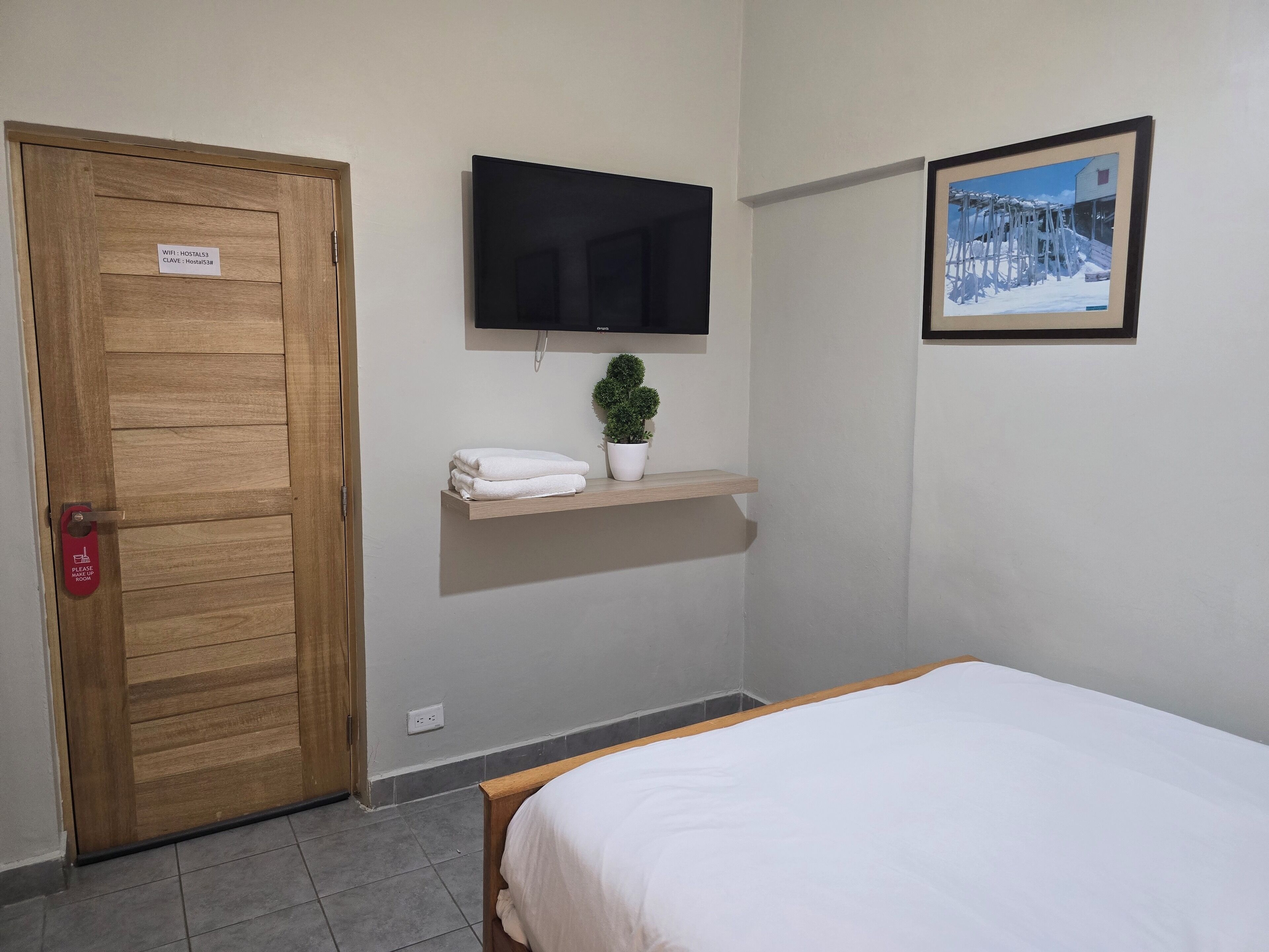 Habitación de hotel con puerta de madera, televisor de pantalla plana en la pared, una planta pequeña en maceta, toalla blanca sobre un estante y un cuadro enmarcado en la pared.
