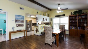 A spacious kitchen with wooden flooring, a dining area with a wicker chair, and a bookshelf filled with books and decorative items.