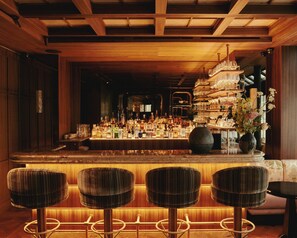 A bar area with a wooden ceiling, a variety of bottles, glasses, and a vase with flowers. There are four wicker bar stools in front. - Hotel Hana (Paris)