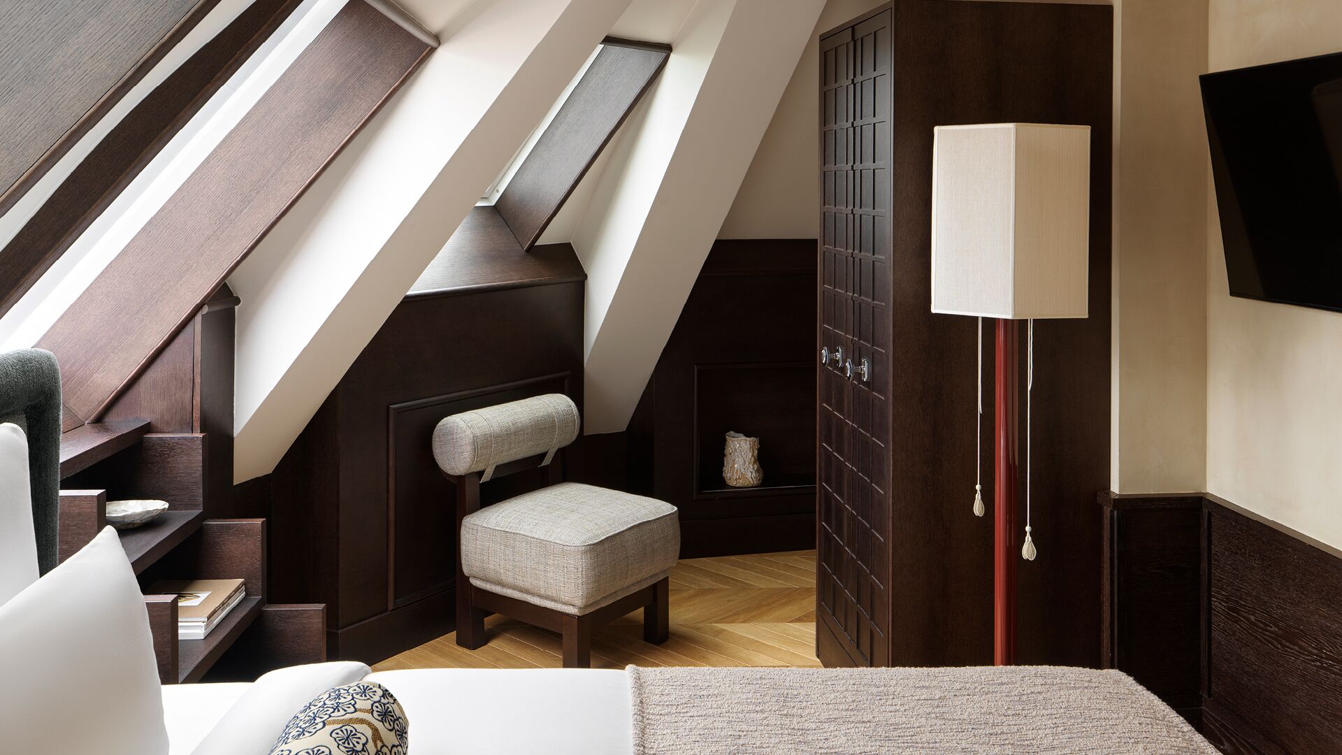 A bedroom with a sloped ceiling, a chair, a bedside table, a lamp, and a wardrobe.
