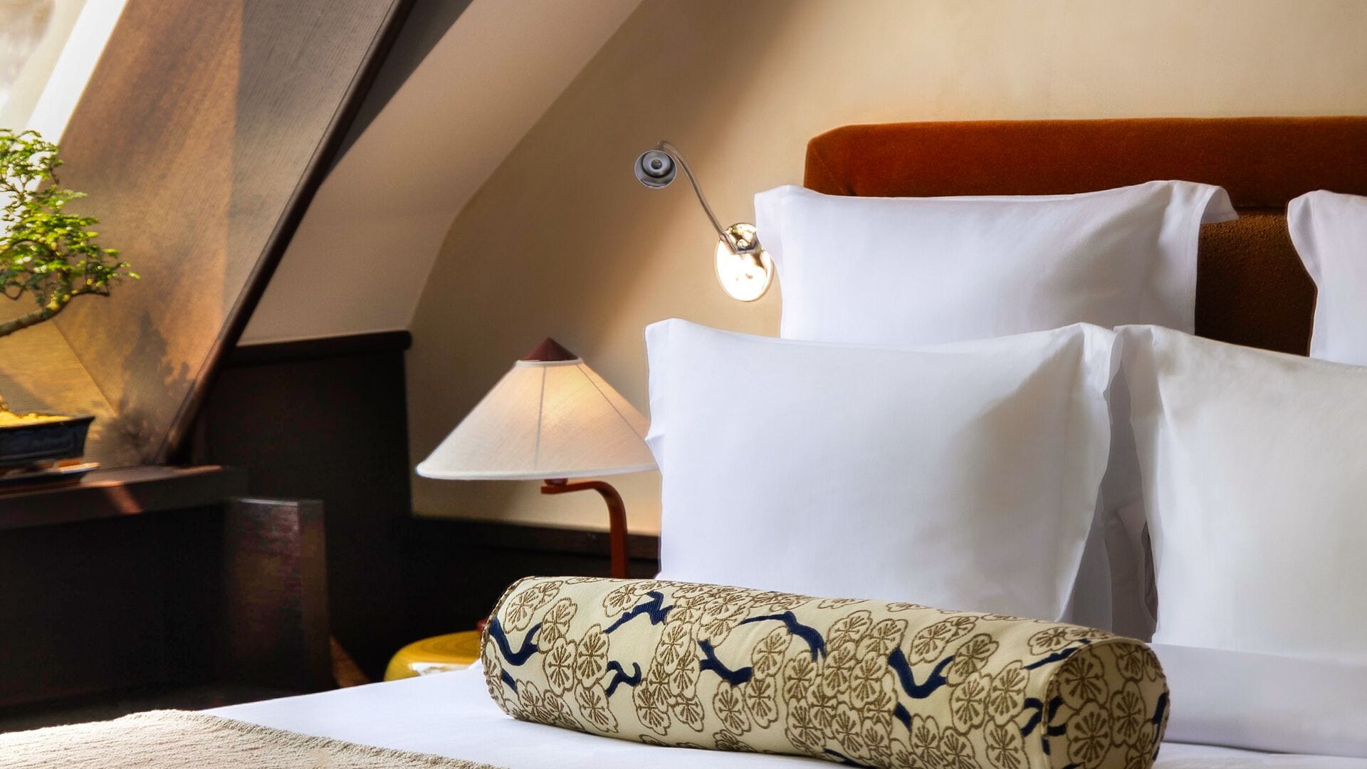 Una cama bien tendida con sábanas blancas, un cojín decorativo, una lámpara de noche y una mesita de noche con una planta.