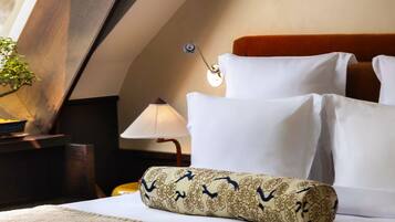Una cama bien tendida con sábanas blancas, un cojín decorativo, una lámpara de noche y una mesita de noche con una planta.