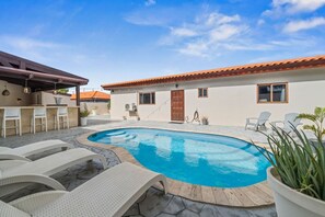 A swimming pool with lounge chairs, a covered patio area, and a building with a tiled roof. - Felicidad: Stunning 3BR Retreat with Private Pool (Oranjestad)
