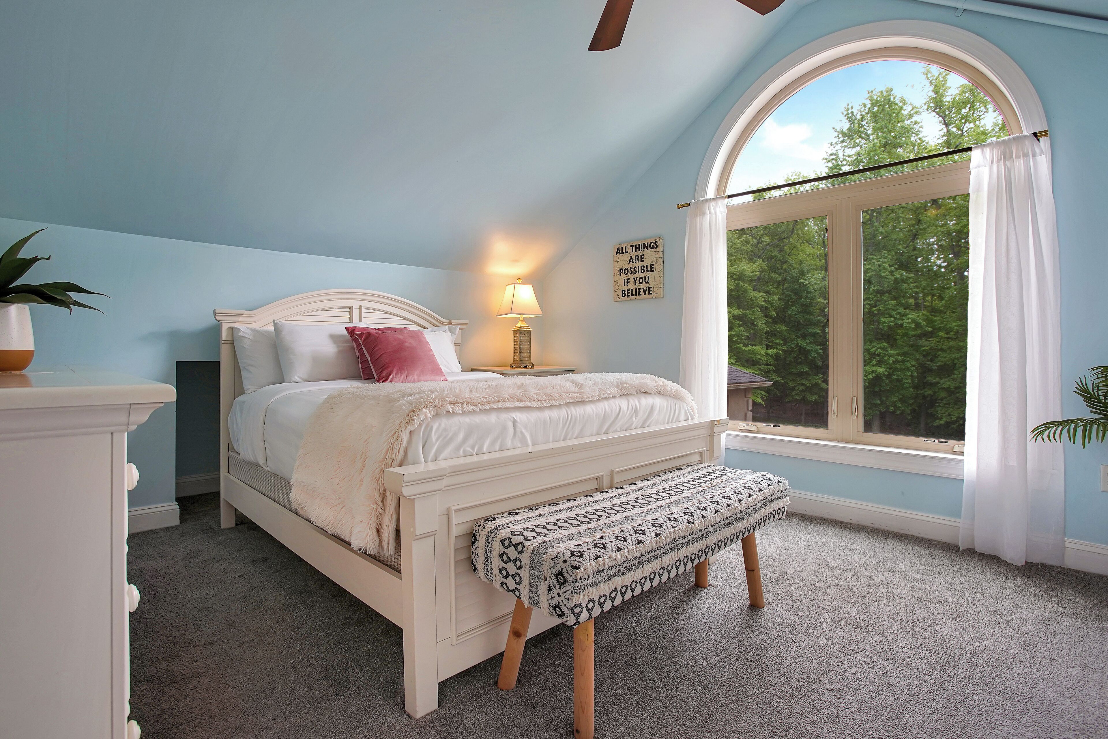 A bedroom with a bed, bedside table, lamp, bench, and a window with curtains.