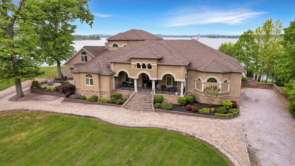 A large, two-story house with a stone facade, a covered patio, and a view of a lake.