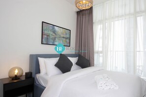 Uma cama bem arrumada com lençóis brancos e um conjunto de toalhas, um abajur, um quadro abstrato na parede e uma janela grande com cortinas leves.