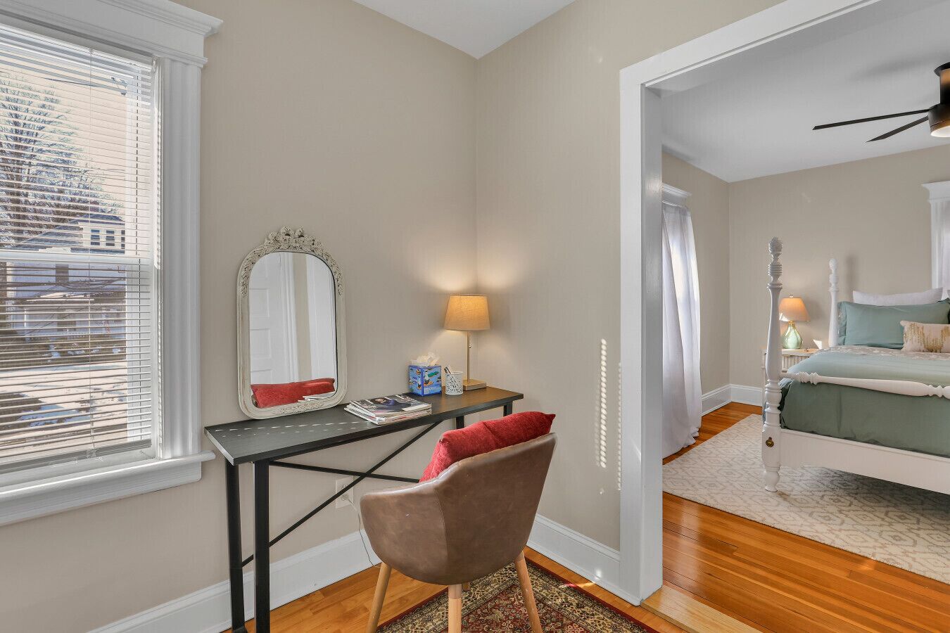 Une chambre à coucher avec un lit, un bureau avec une chaise, un miroir et une fenêtre avec des stores.