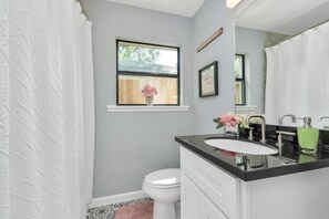Un baño con inodoro blanco, lavamanos negro, espejo y ventana con marco de madera.