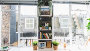 A room with a large window, a bookshelf with books and decorative items, a small table with a bottle and glasses, and a fan on the windowsill. - Stylish & cozy City Studio close to Hyde Park - Pass the Keys (London)