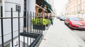 A street view with a building featuring a black metal fence, a red car parked on the side, and a green awning over the entrance. - Stylish & cozy City Studio close to Hyde Park - Pass the Keys (London)