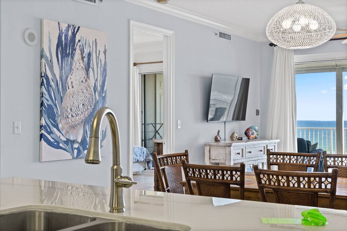 A modern kitchen with a large island, a dining area with wooden chairs, a white cabinet, and a large window with a view of the ocean.