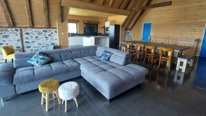 Una sala moderna con un sofá seccional gris, un otomano pequeño y redondo, y una silla amarilla. Se ve un comedor con una mesa y sillas de madera al fondo.
