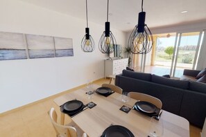 Un espace repas moderne avec une table en bois dressée pour quatre personnes, un canapé gris et une vue sur l’extérieur grâce à de grandes fenêtres.