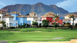 A golf course with a row of colorful villas, each with a distinct architectural style and a mountain backdrop.