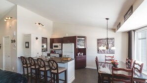 A modern kitchen with a central island, dining area with wooden chairs, and a large window providing natural light. - Le Bionick- Condo à louer Stoneham  (Stoneham-et-Tewkesbury)