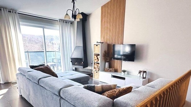 A modern living room with a grey sectional sofa, a flat-screen TV mounted on the wall, and a large window with curtains. - Le Bionick- Condo à louer Stoneham  (Stoneham-et-Tewkesbury)