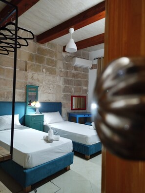 A room with two beds, a stone wall, a wooden ceiling, a white air conditioner, and a blue bedside table with a lamp. - Limestone Sun Suites (Sliema)