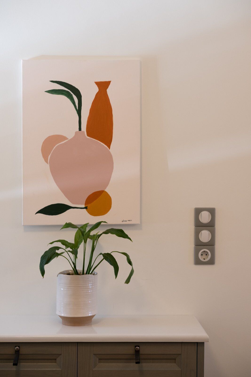 A wall-mounted artwork depicting abstract shapes in orange and pink, a potted plant on a white surface, and a wall socket with three switches.
