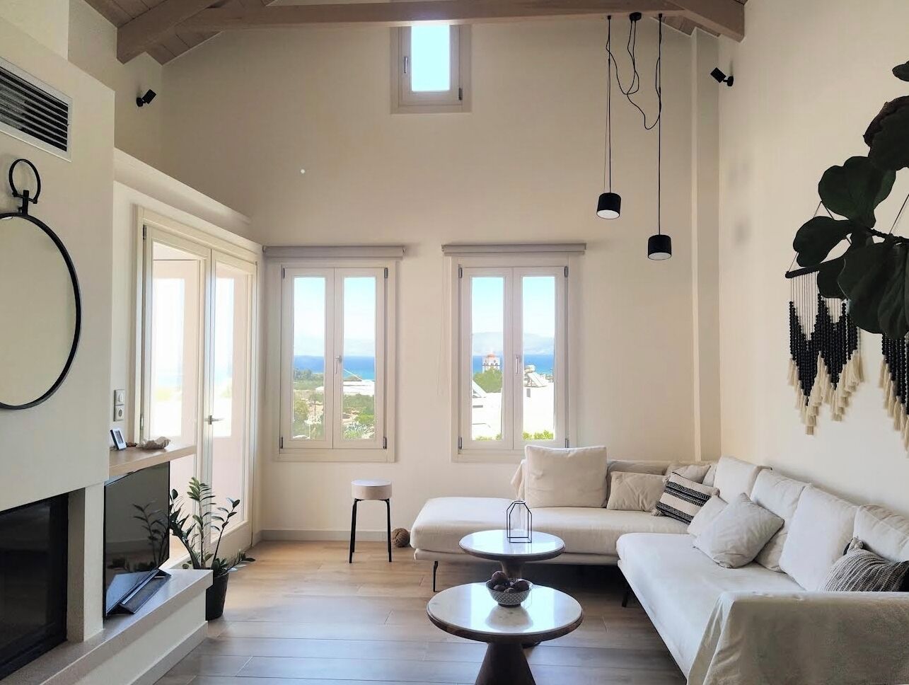 Ein geräumiges Wohnzimmer mit einem weißen Sofa, einem runden Couchtisch und einem Kamin. Große Fenster bieten einen Blick ins Freie.
