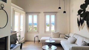 A spacious living room with a white sofa, a round coffee table, and a fireplace. There are large windows providing a view of the outdoors.