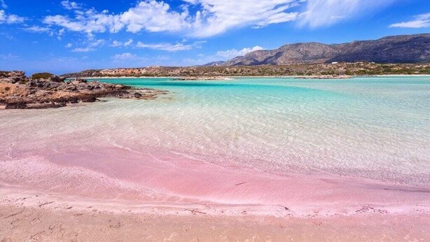 A beach with clear turquoise water, a pink sand foreground, and rocky hills in the background.
