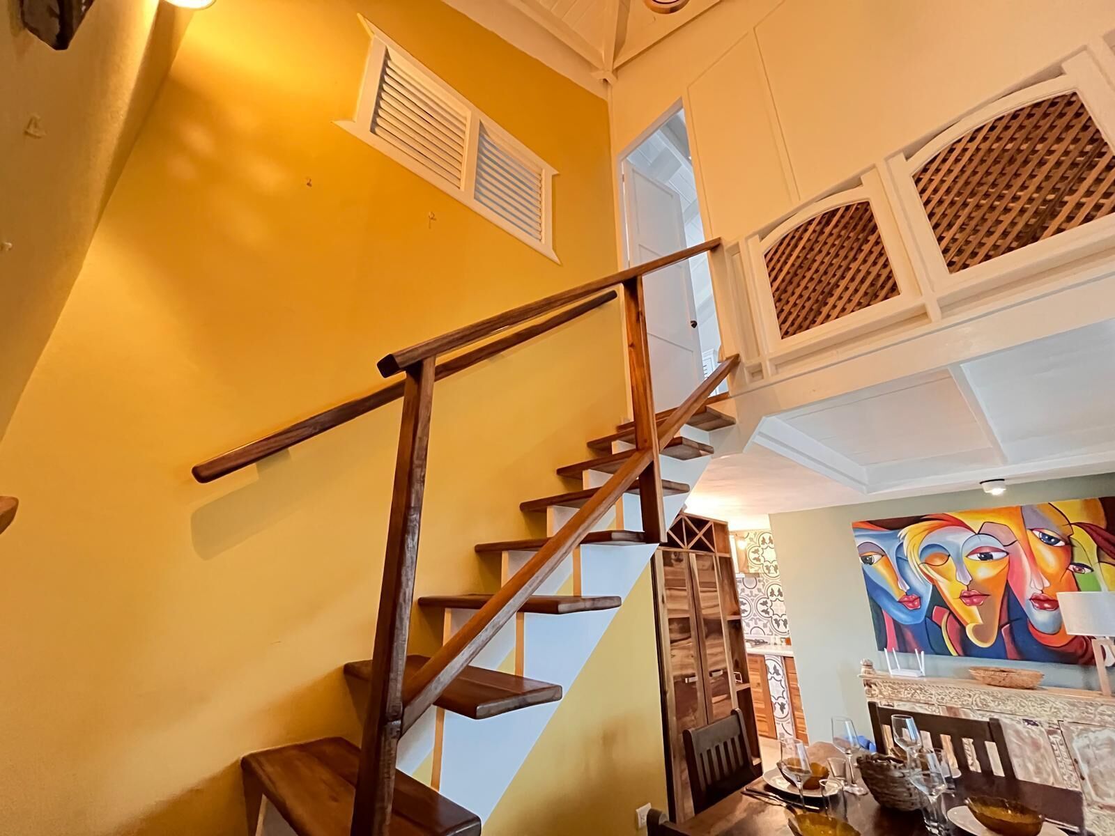Una escalera con barandillas de madera, un comedor con una mesa dispuesta para dos personas y un cuadro colorido en la pared.