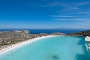 A clear blue sky, a curved swimming pool, and a beach with rocky terrain.
