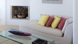 Una sala de estar moderna con una mesa de centro de vidrio, un sofá blanco con cojines estampados, una chimenea con objetos decorativos y una obra de arte enmarcada en la pared.