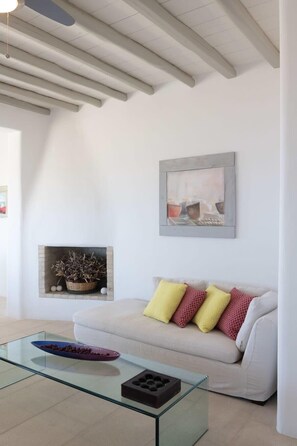 A modern living room with a glass coffee table, a white sofa with patterned cushions, a fireplace with decorative items, and a framed artwork on the wall.