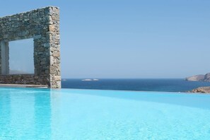 Una estructura de arco de piedra sobre una piscina azul cristalina, con vistas al mar y a la costa lejana.