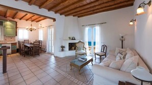 A spacious living area with a wooden ceiling, a dining table, a sofa, and a fireplace. - Villa Victoria with Sea View and Two Private Pools (Mykonos)