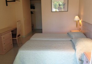 Quarto com duas camas, uma escrivaninha, uma cadeira e uma mesa de cabeceira com abajur.