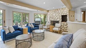 A spacious living room with a stone fireplace, large windows, and a modern kitchen area.