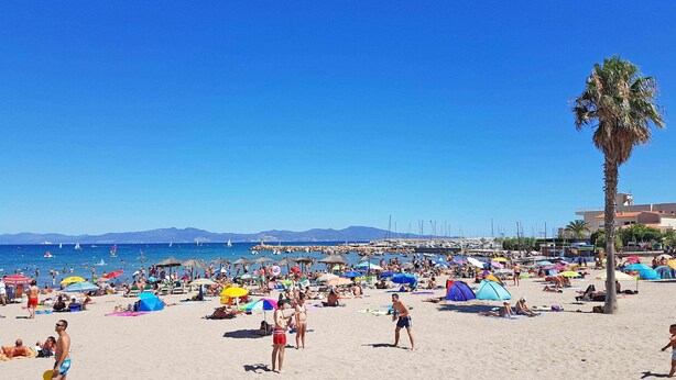 Ein überfüllter Strand mit vielen Sonnenschirmen und Zelten, einer Palme und Booten im Hintergrund.