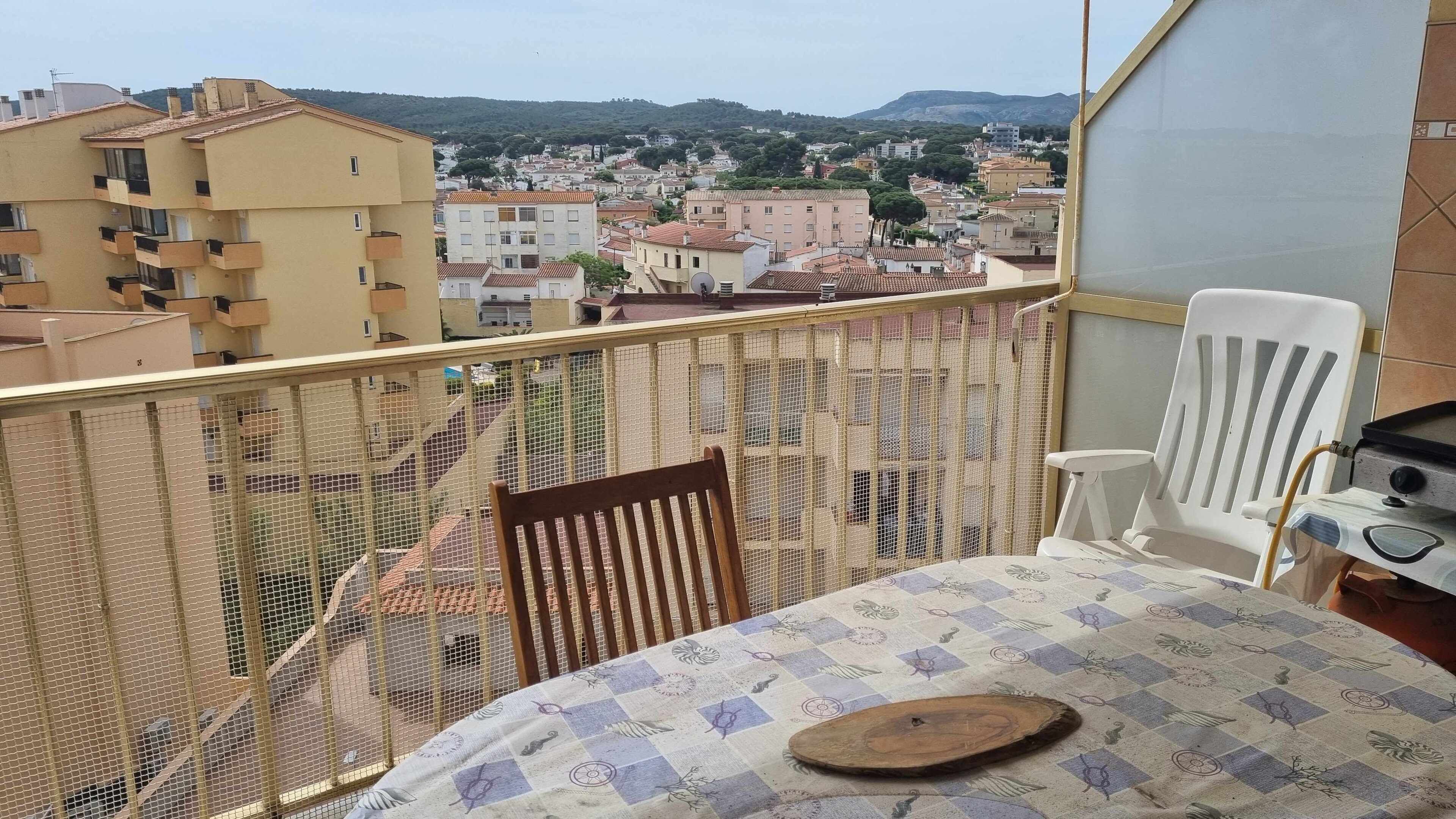 Un balcón con vista a un pueblo, una silla blanca, una mesa con mantel estampado y una bandeja de madera.